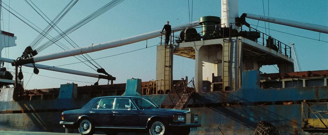 Lord of War (2005) movie still: wide — A dark sedan sits on a dock next to a large, rusted cargo ship. A lone man stands on the upper steel deck of…