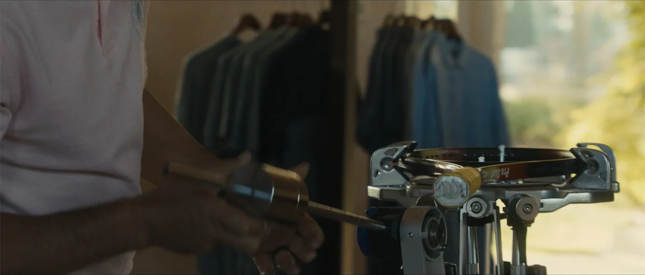 King Richard (2021) movie still: medium — A well-lit room displays racks of clothing on hangers in the soft-focus background. A man uses a metal…