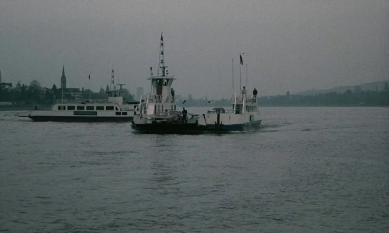 The Lost Honour of Katharina Blum (1975) movie still: wide — Two small car ferries navigate a wide, calm river under an overcast, grey sky. Distant…