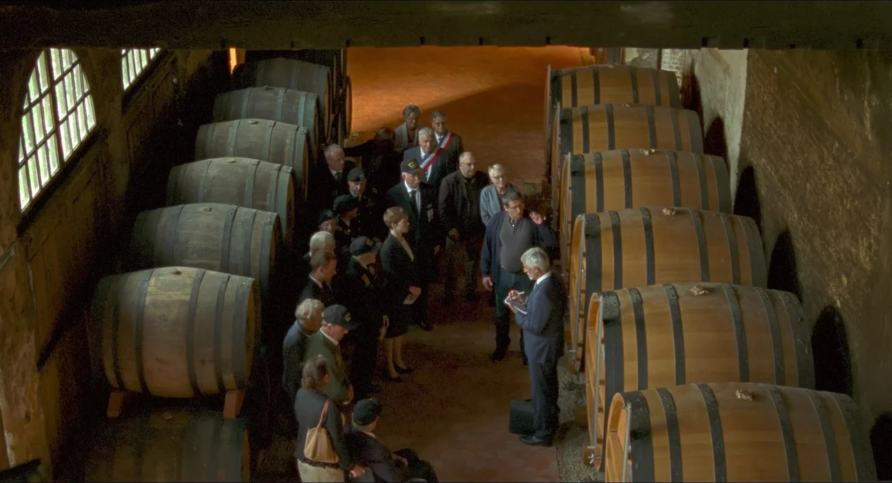 One Fine Morning (2022) movie still: wide — An interior wine cellar features two long rows of large stacked oak barrels. A diverse group of people…