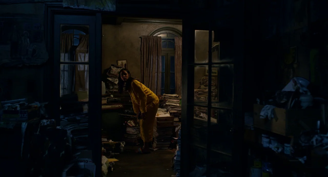 Velvet Buzzsaw (2019) movie still: fullbody — A dark room is filled with overflowing stacks of papers and boxes. A woman in yellow pajamas leans…