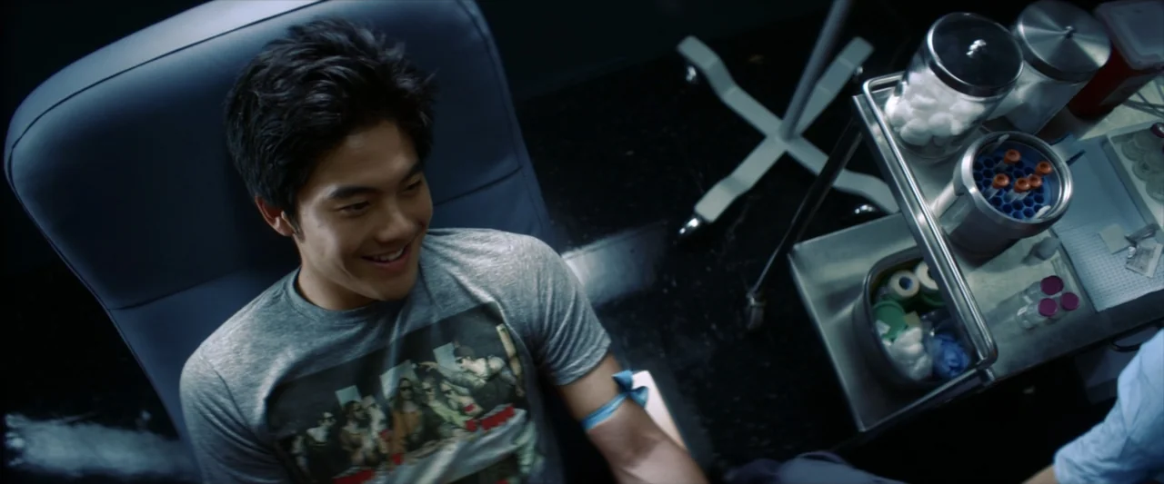 Tell Me How I Die (2016) movie still: medium — A clinical medical space features a phlebotomy chair and a rolling metal supply cart. A young man…