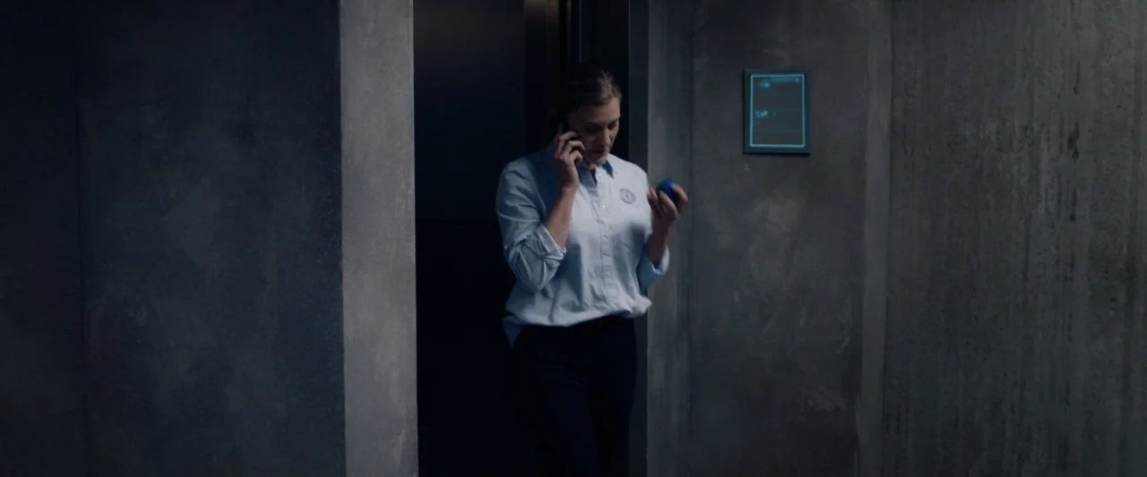 2036 Origin Unknown (2018) movie still: medium — A woman exits a doorway into a narrow corridor featuring bare grey concrete walls. She holds a…