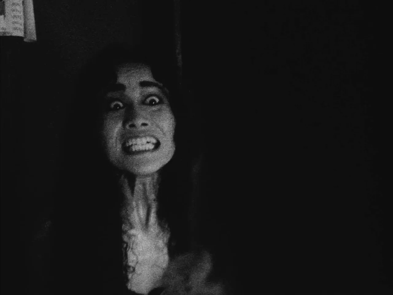 Tetsuo: The Iron Man (1989) movie still: closeup — A dark, dimly lit room provides no background detail. A woman stares into the camera with her…