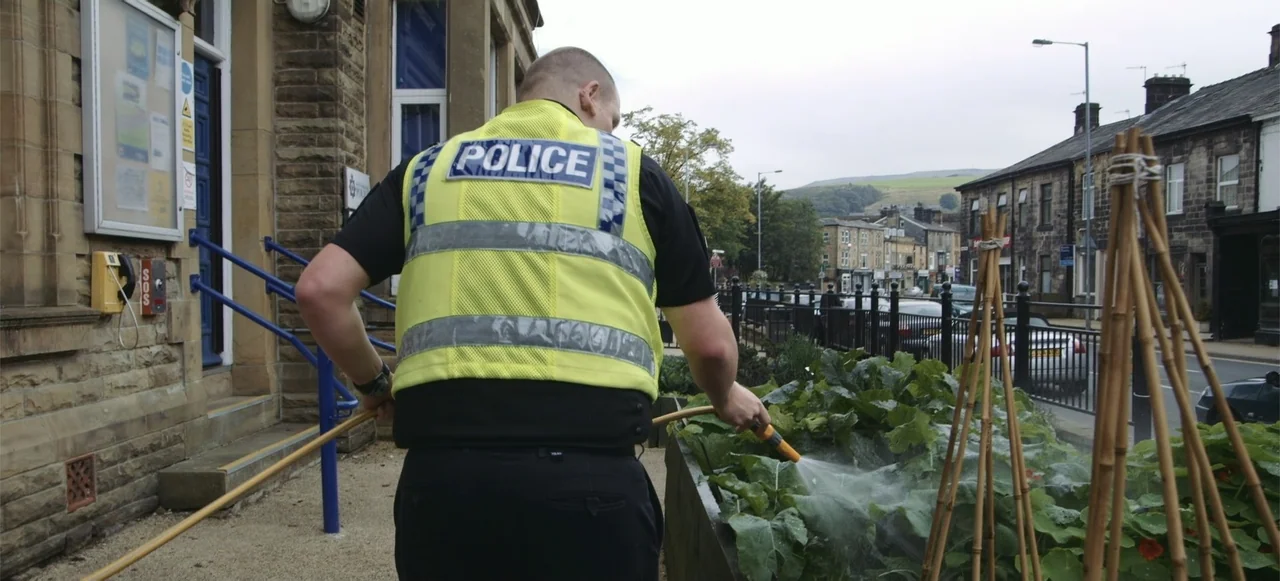10 Billion: What's On Your Plate (2015) movie still: medium — A public sidewalk area adjacent to a stone building with a small garden bed. A police…