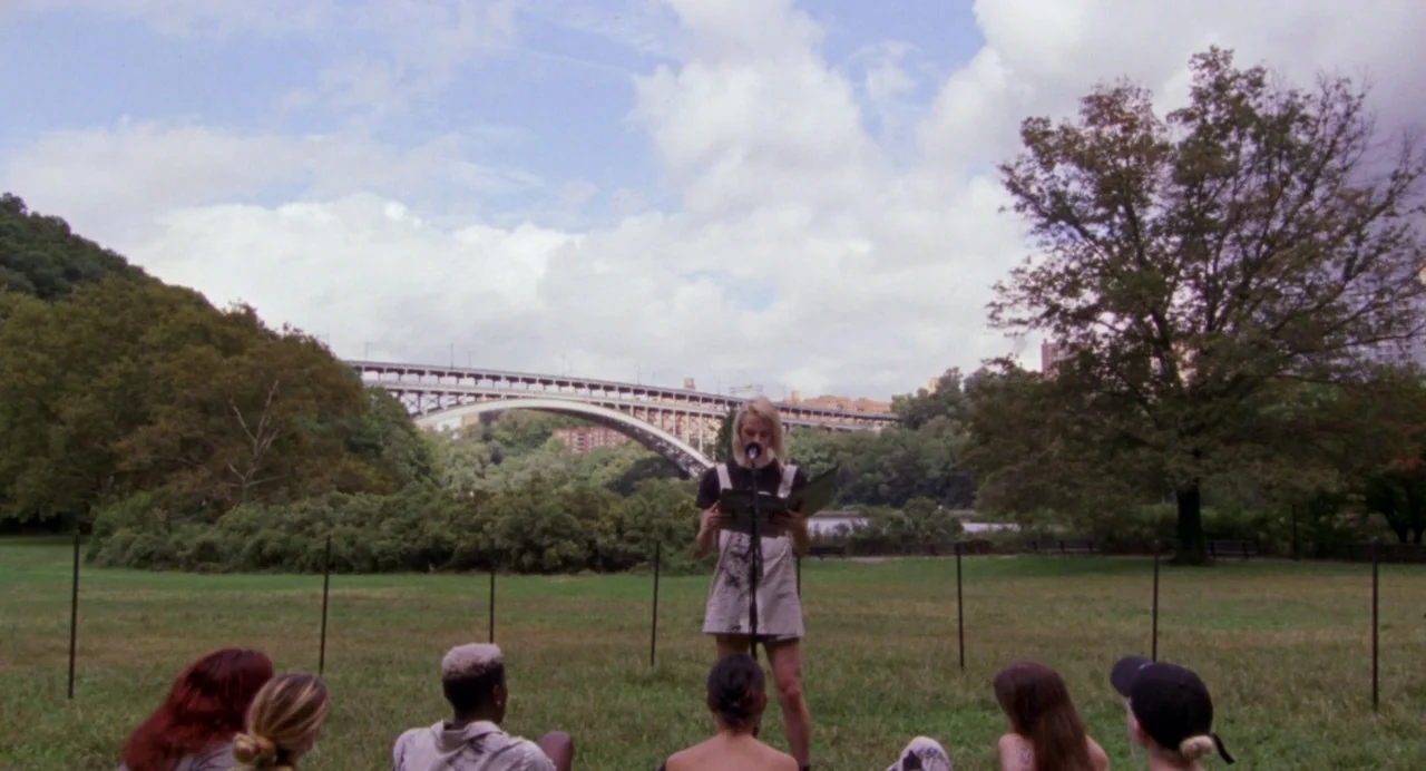 So Pretty (2019) movie still: wide — A green grassy park space with a large arch bridge visible in the background. A woman stands alone at a…