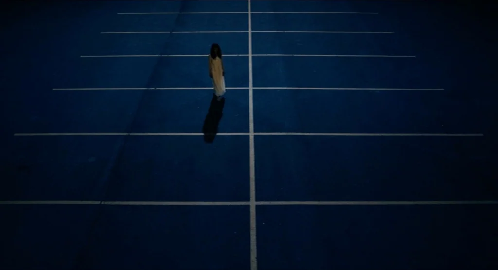 Life During Wartime (2009) movie still: wide — A dark, empty asphalt parking lot is marked by stark white painted lines. A lone woman in a white…