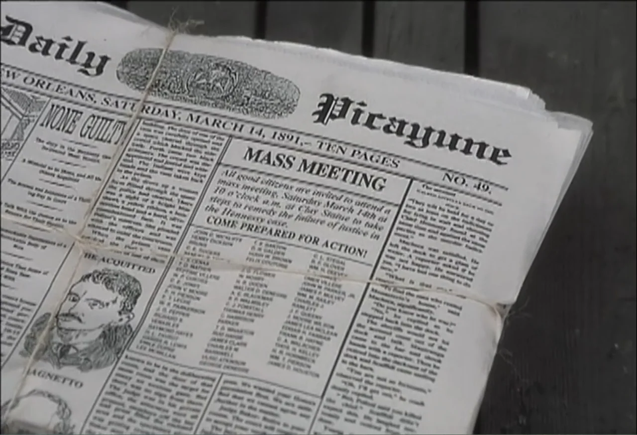 Vendetta (2022) movie still: closeup — An aged newspaper dated 1891 lies flat on a dark wooden plank surface. The paper is folded and secured with a…