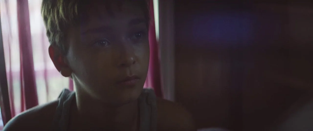 The Strange Ones (2017) movie still: closeup — An interior bedroom space with thin purple curtains visible in the background near a window. A boy…