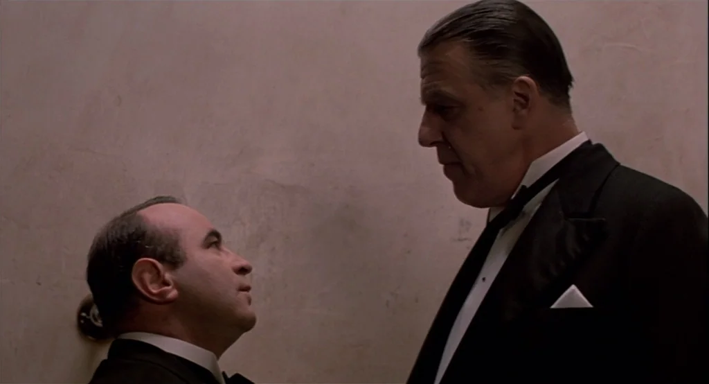 The Cotton Club (1984) movie still: medium — A dimly lit, narrow stairwell with textured light-colored walls houses two men. A short man and a tall…