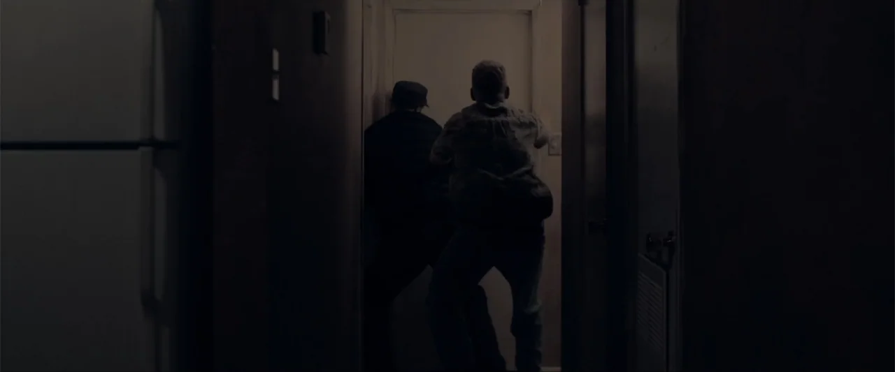 Anomaly (2006) movie still: fullbody — A dimly lit narrow residential hallway leads toward a light-colored closed door. Two individuals stand in the…