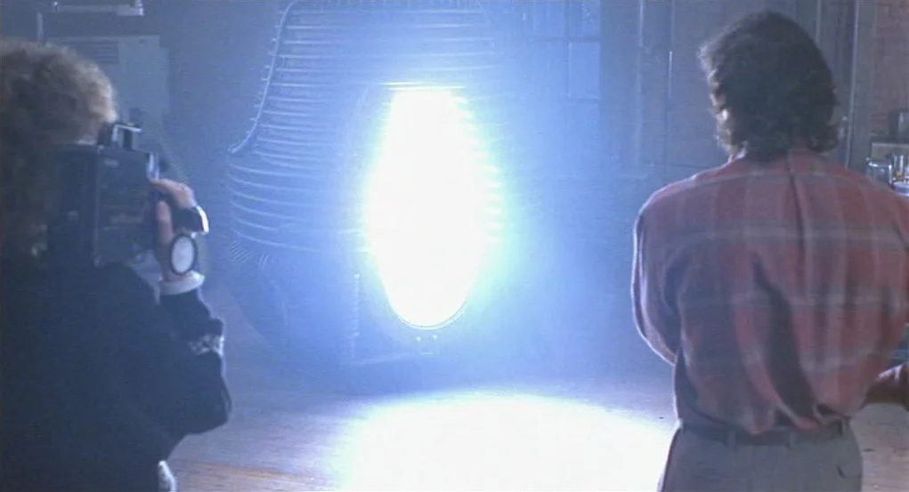 The Fly (1986) movie still: medium — A dimly lit basement features a glowing metallic device centered between two people. One person holds a camera…