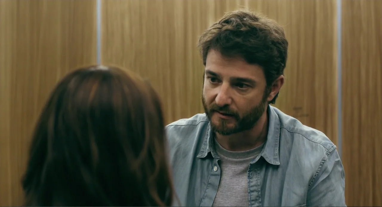 The Lift (2021) movie still: medium — The interior of an elevator is paneled with light brown vertical wood planks. A bearded man in a denim shirt…
