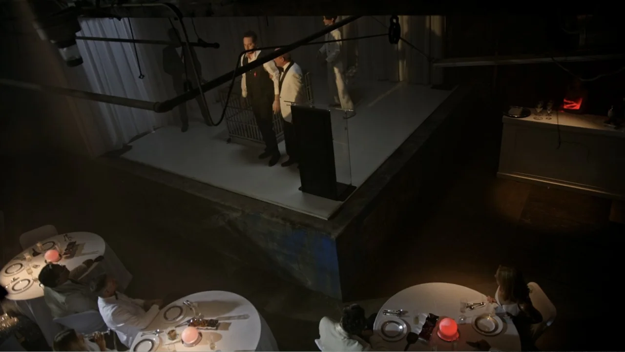 The Collective (2023) movie still: wide — A dimly lit room contains a raised white stage platform and several circular tables. Guests sit at the…