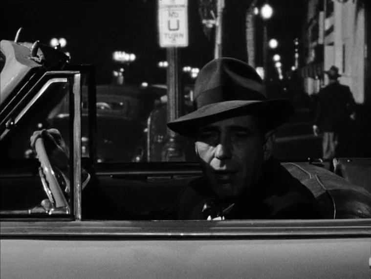 In a Lonely Place (1950) movie still: medium — A man sits in the driver's seat of an open convertible at night. He is steering the vehicle while…