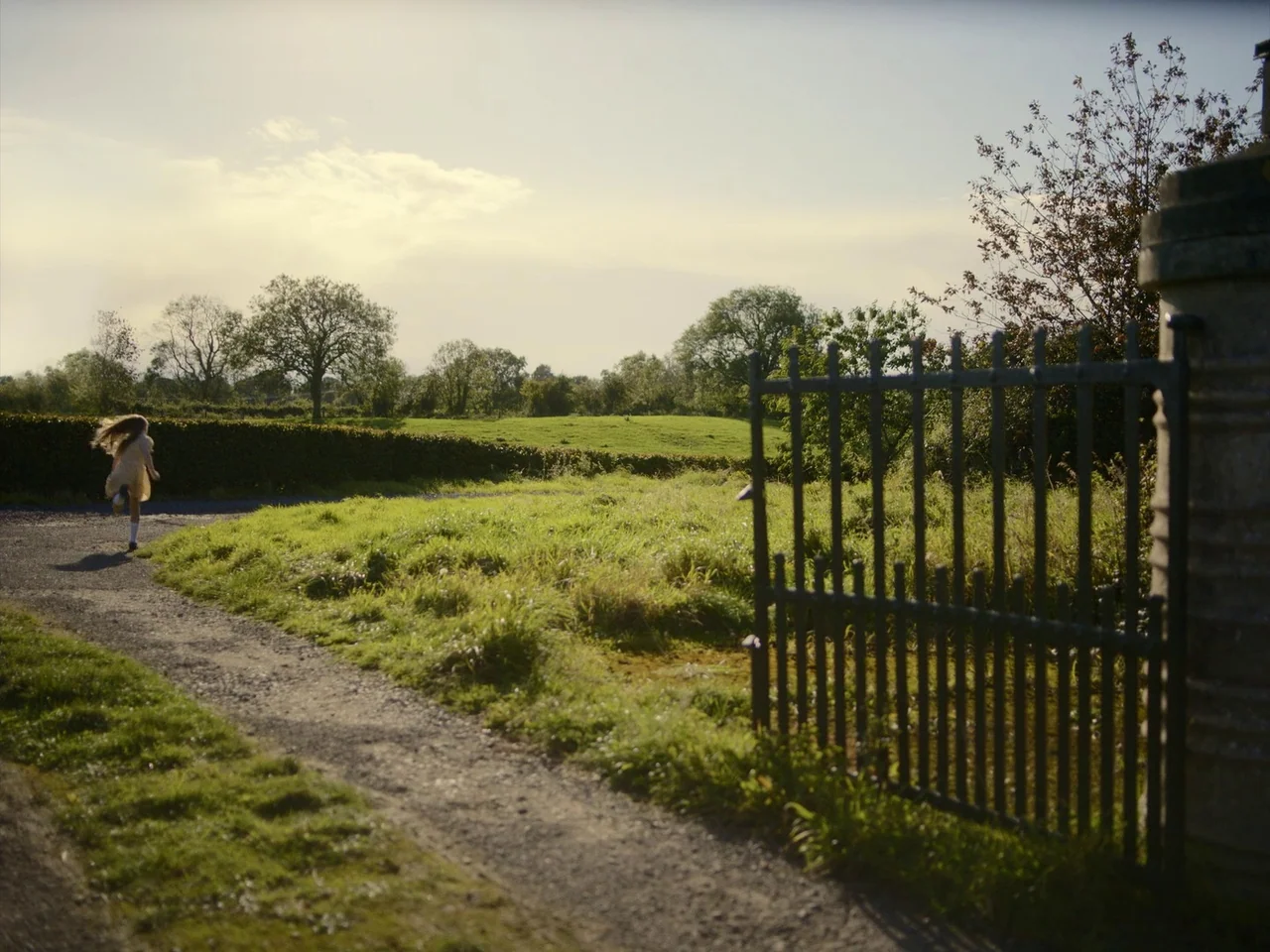 The Quiet Girl (2022) movie still: wide — A gravel path curves through a grassy rural landscape bordered by hedges and trees. A young girl in a pale…