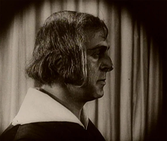 Destiny (1921) movie still: closeup — A close-up view shows a man standing in front of heavy fabric curtains. The subject appears in profile while…