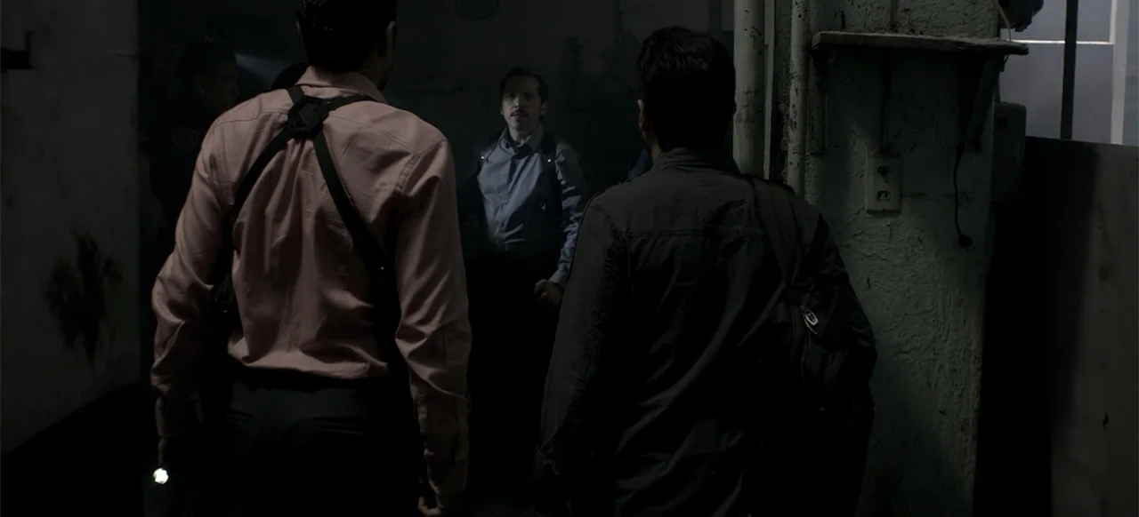 About the Deaths (2016) movie still: medium — A dimly lit narrow hallway with textured walls features two men standing in the foreground. A third…