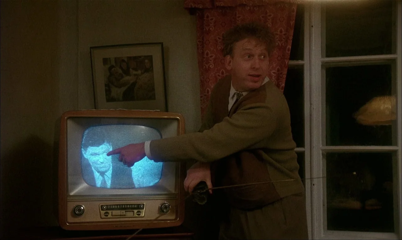 My Life As A Dog (1985) movie still: medium — A man stands in a dimly lit room beside a vintage wooden television cabinet. He points his finger at a…
