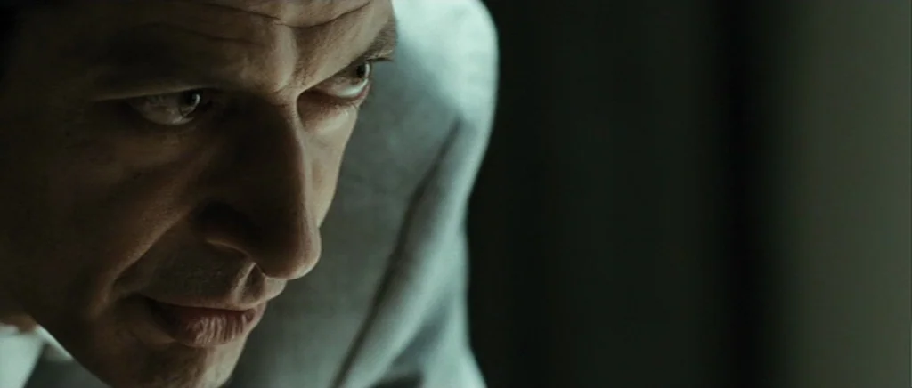 Adam Resurrected (2008) movie still: closeup — A dimly lit room with a man in a grey suit occupies the frame. The subject leans forward with his…