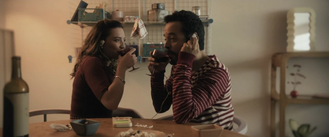 Sign Here (2017) movie still: medium — A light-toned interior space features a small wooden table with plates and glasses. A man and woman sit…