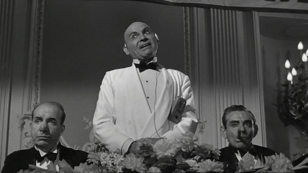 Some Like It Hot (1959) movie still: medium — Three men in formal dinner attire sit at a head table adorned with flowers. The central figure wears…