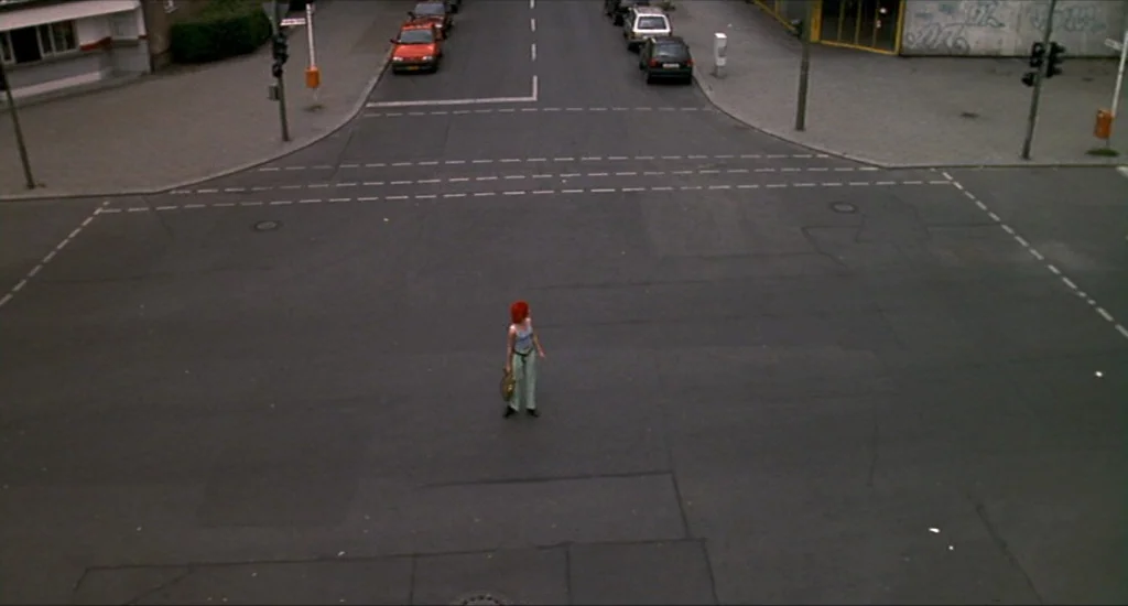 Run Lola Run (1998) movie still: wide — A wide paved intersection in a European urban area contains road markings and traffic signals. A woman with…