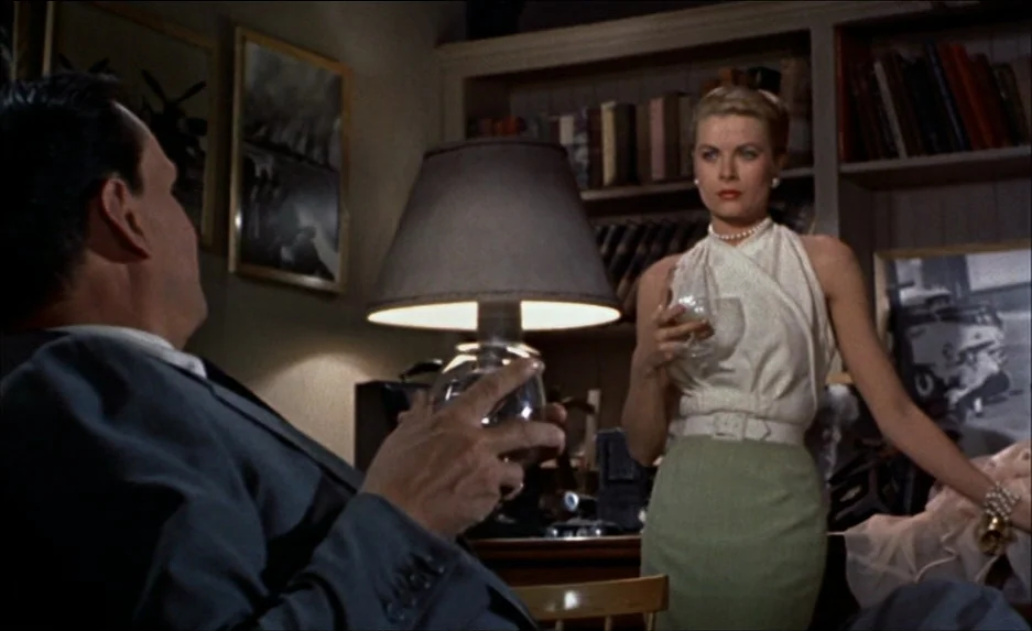 Rear Window (1954) movie still: medium — A dark study features wooden bookshelves and framed pictures on the wall behind. A woman holding a glass…
