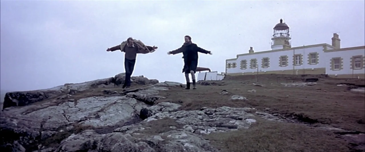 Breaking the Waves (1996) movie still: wide — A grassy cliff top features a white painted lighthouse building with multiple square windows. A man…