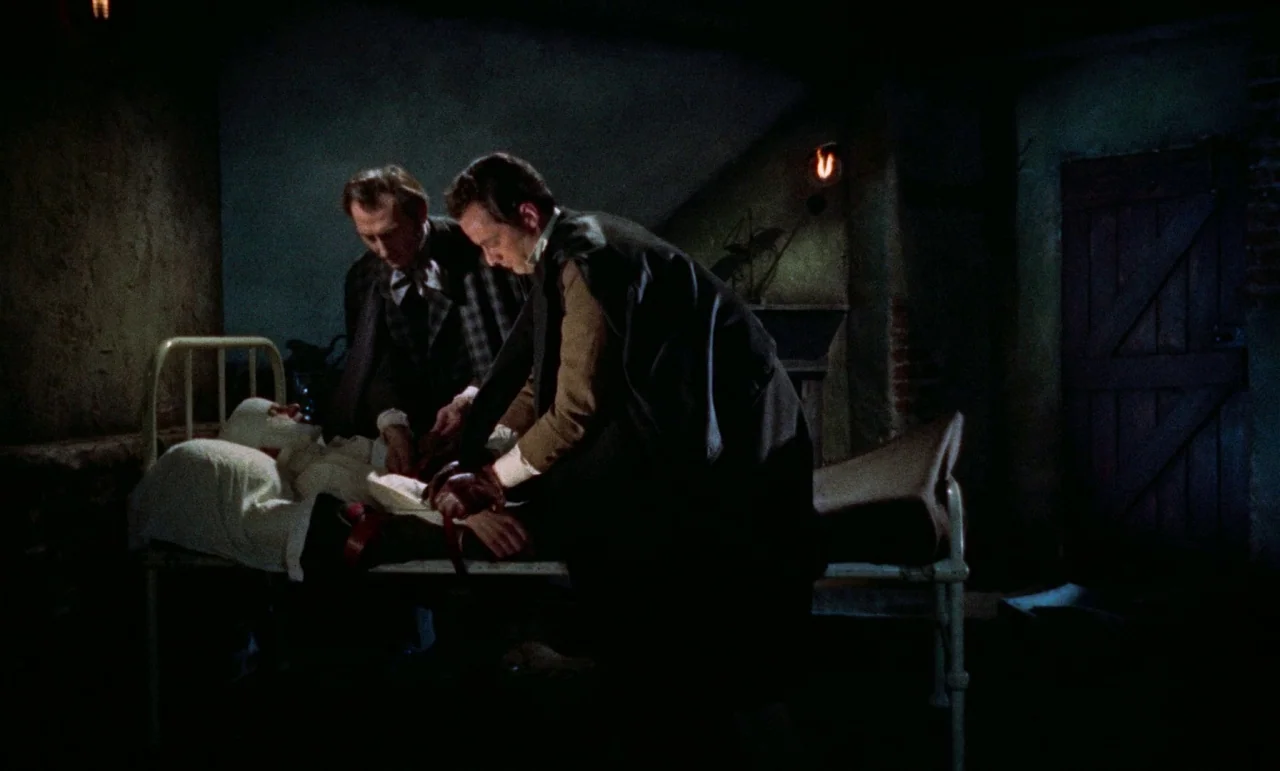 The Revenge of Frankenstein movie still: medium — A dimly lit room contains a white metal hospital bed and a wooden door. Two men inspect the…