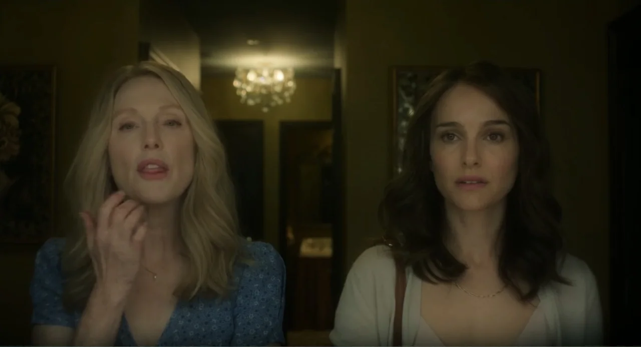 May December (2023) movie still: medium — A dimly lit indoor residential corridor with dark walls and a glowing chandelier. Two women stand…