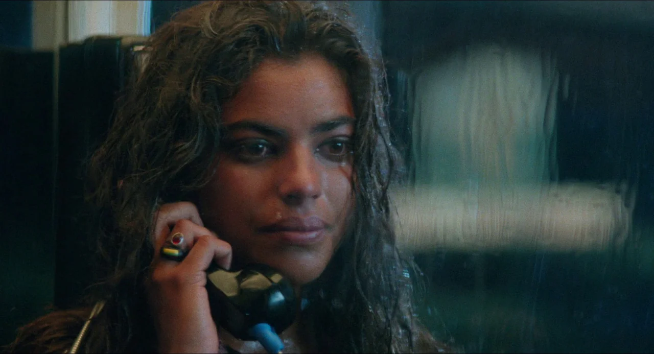 Mississippi Masala (1991) movie still: closeup — A dark phone booth interior with condensation on the glass pane. A woman with wet hair listens to a…