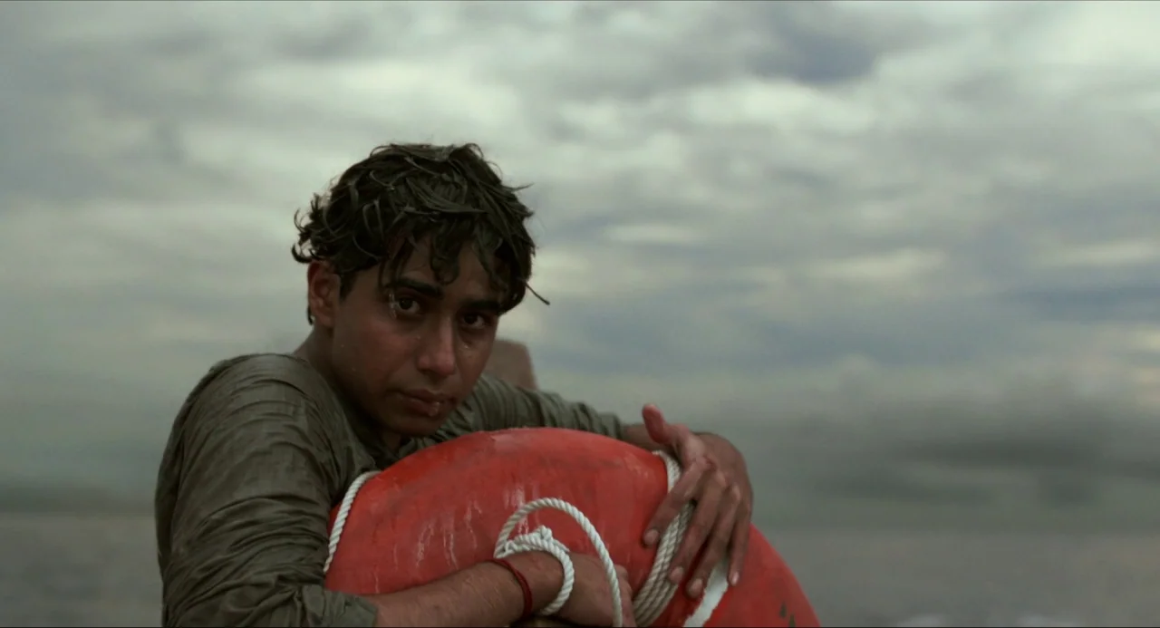Life of Pi (2012) movie still: medium — An overcast sky hangs over a vast expanse of gray, choppy ocean water. A young man sits in the water…