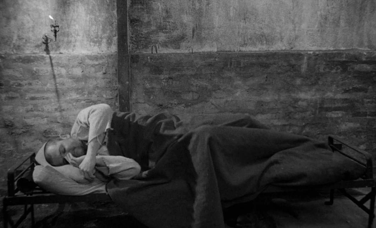 Goto, Island of Love (1969) movie still: fullbody — A austere stone-walled enclosure features a thin metal cot and a single wall sconce. A man lies…