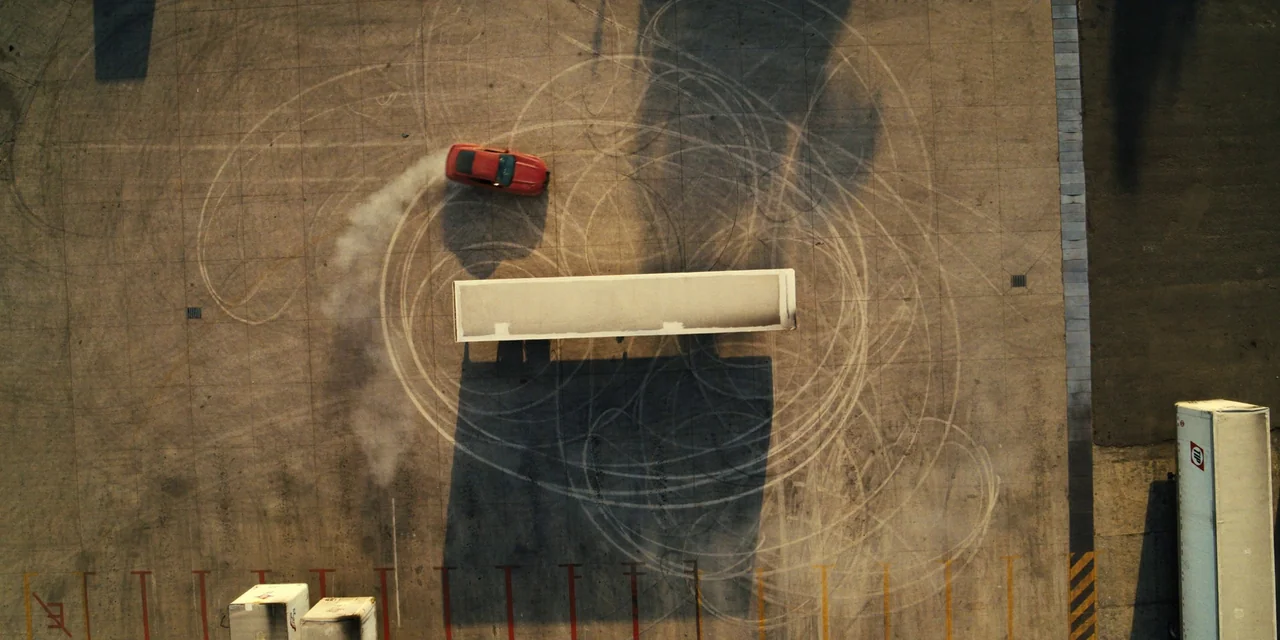 Silent Night (2023) movie still: wide — A wide asphalt parking lot is marked by multiple circular rubber tire tracks. A red sports car performs a…
