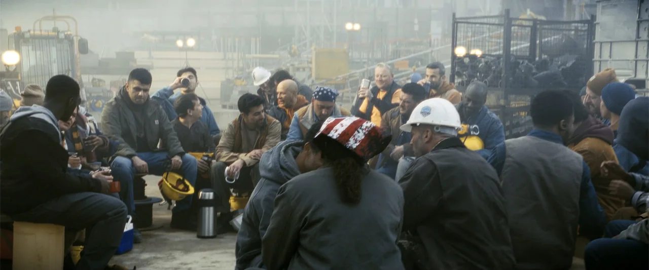 A Working Man (2025) movie still: wide — A dusty construction site features a gathering of workers huddled in a large circle. The laborers wear work…