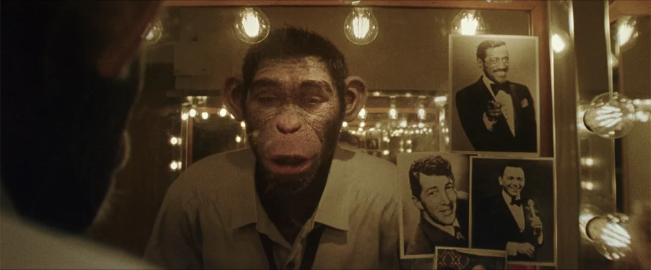 Better Man (2024) movie still: closeup — A dimly lit dressing room features a mirror surrounded by glowing round light bulbs. A chimpanzee wearing a…