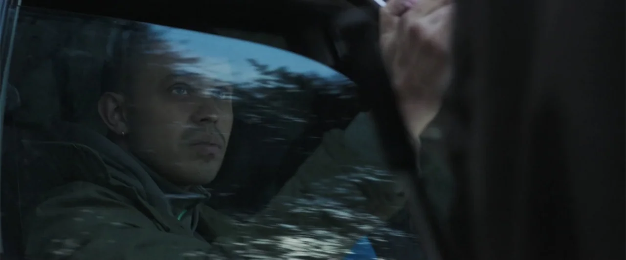 Bring Them Down (2025) movie still: closeup — A person sits inside a car during dusk with the exterior reflected in glass. The driver gazes through…