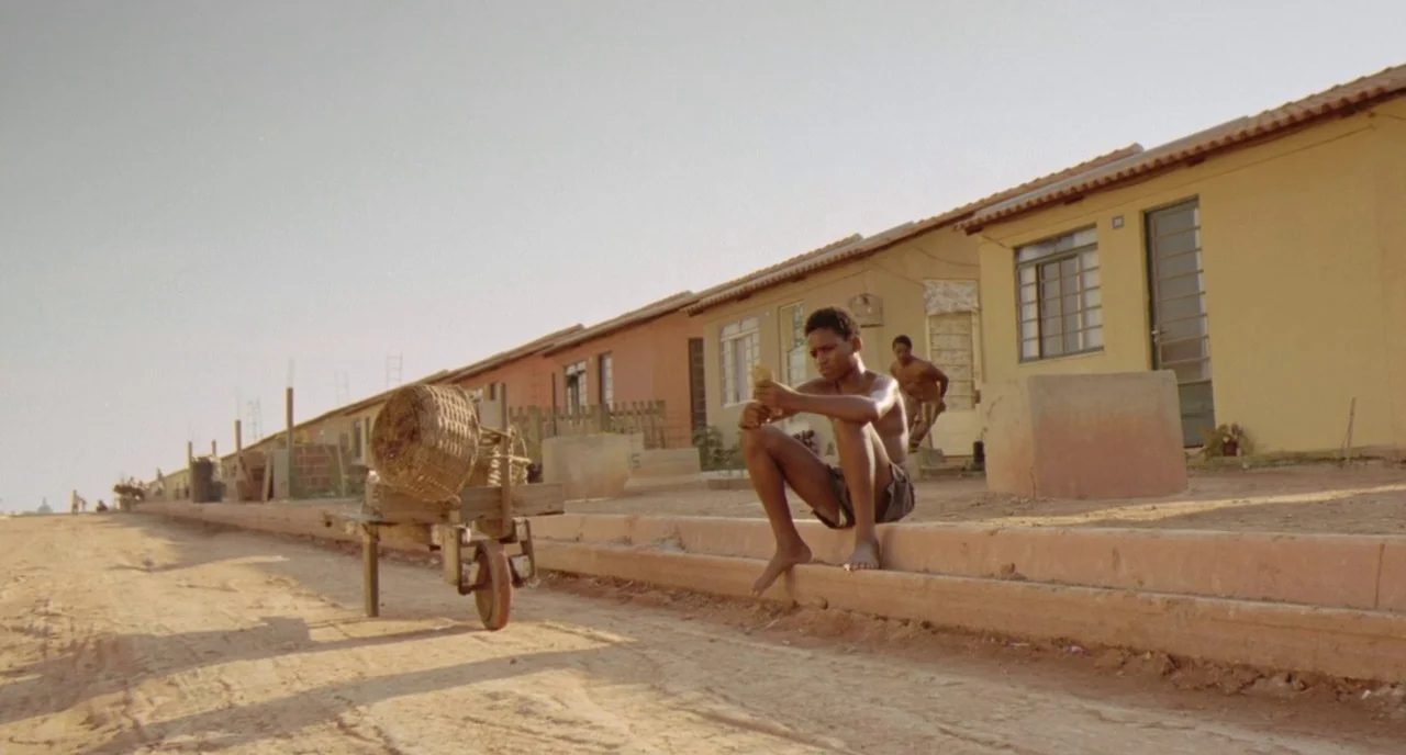 City of God (2002) movie still: fullbody — A dusty, unpaved road borders a row of simple, single-story yellow houses. A young boy sits on the…