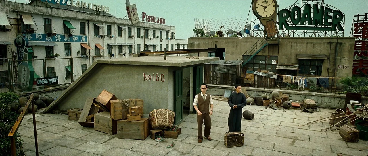 Ip Man 2 (2010) movie still: wide — A cluttered concrete rooftop terrace features wooden crates and baskets in a city setting. Two men stand…