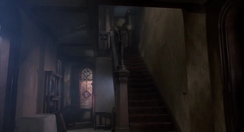Psycho 2 (1983) movie still: establishing — A dark Victorian entrance hall features a wooden staircase and a stained glass window. Dust motes dance…