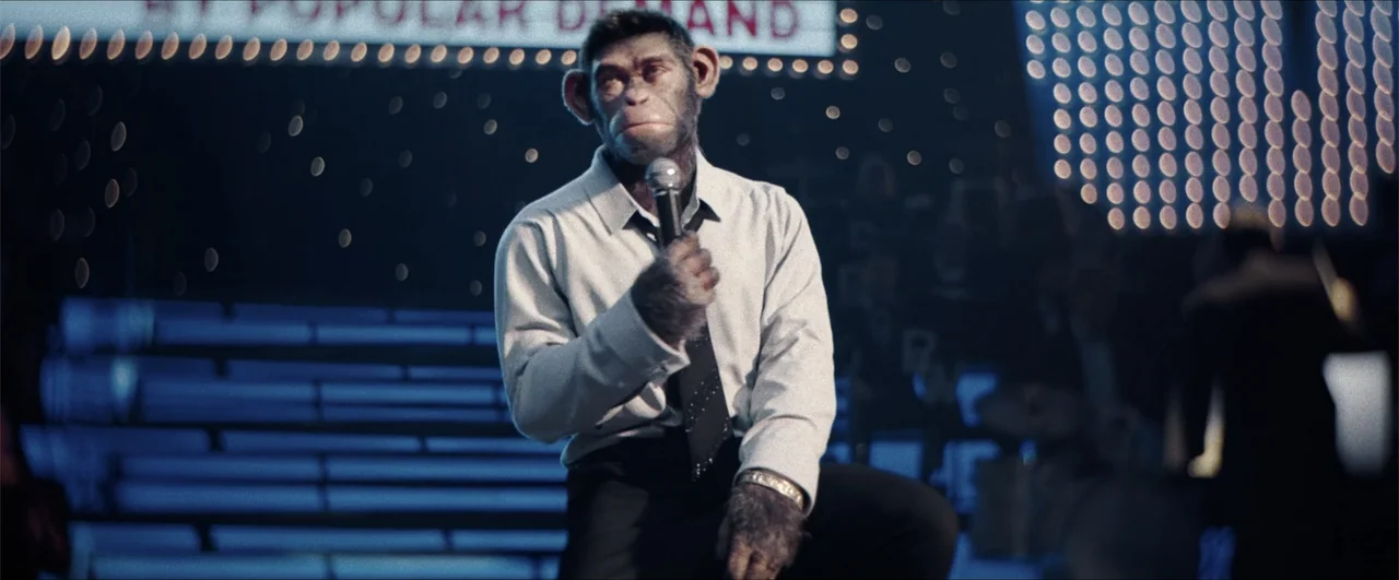 Better Man (2024) movie still: medium — A dimly lit stage features a lone chimpanzee performer before a large illuminated sign. The subject sits on…