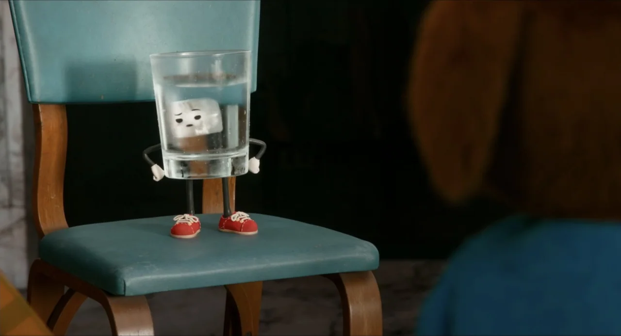 IF (2024) movie still: medium — A teal vinyl chair sits in a room with a blurry out-of-focus foreground subject. An anthropomorphic character made…