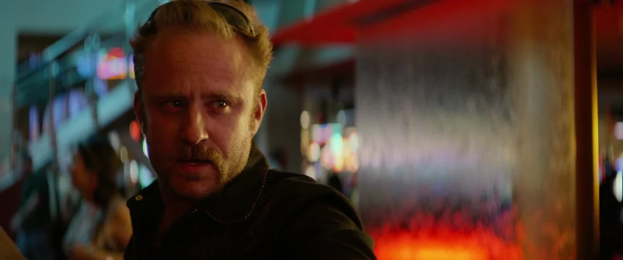 Hell or High Water (2016) movie still: closeup — An interior space of a bar with blurred neon lights in the background. A man with a mustache looks…
