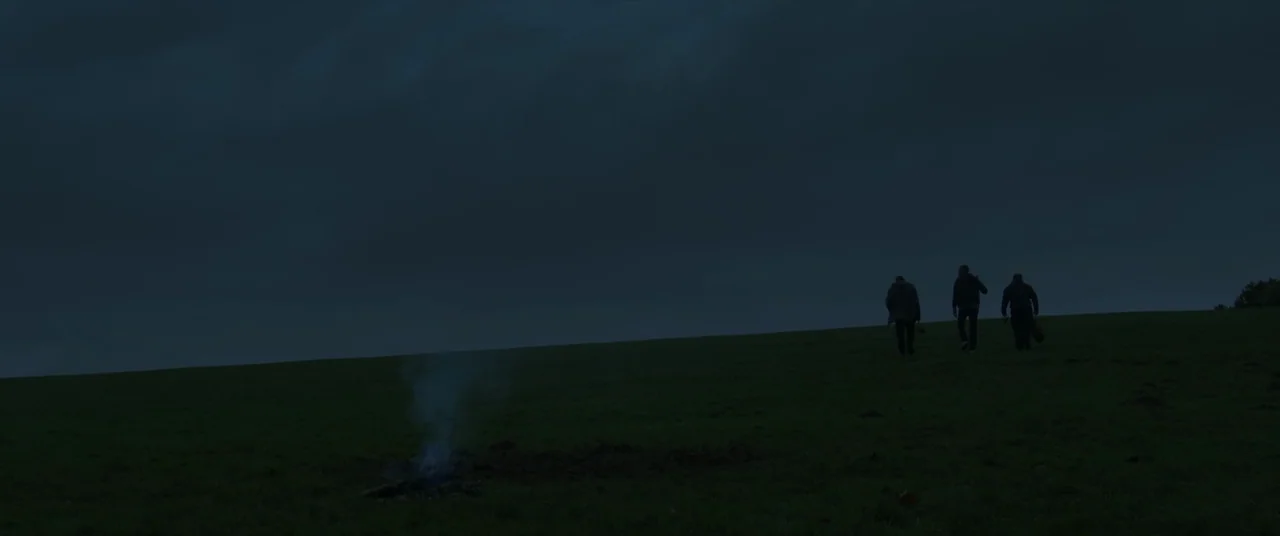 Bull (2021) movie still: wide — A dark, grassy field stretches toward a cloudy night sky under low light. Three figures walk away from a smoldering,…