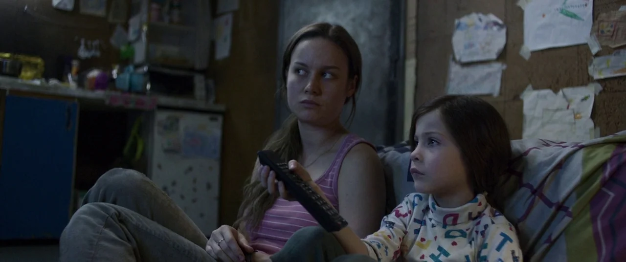 Room (2015) movie still: medium — A dimly lit room features walls covered in taped-up children's drawings and sparse furniture. A young woman and a…