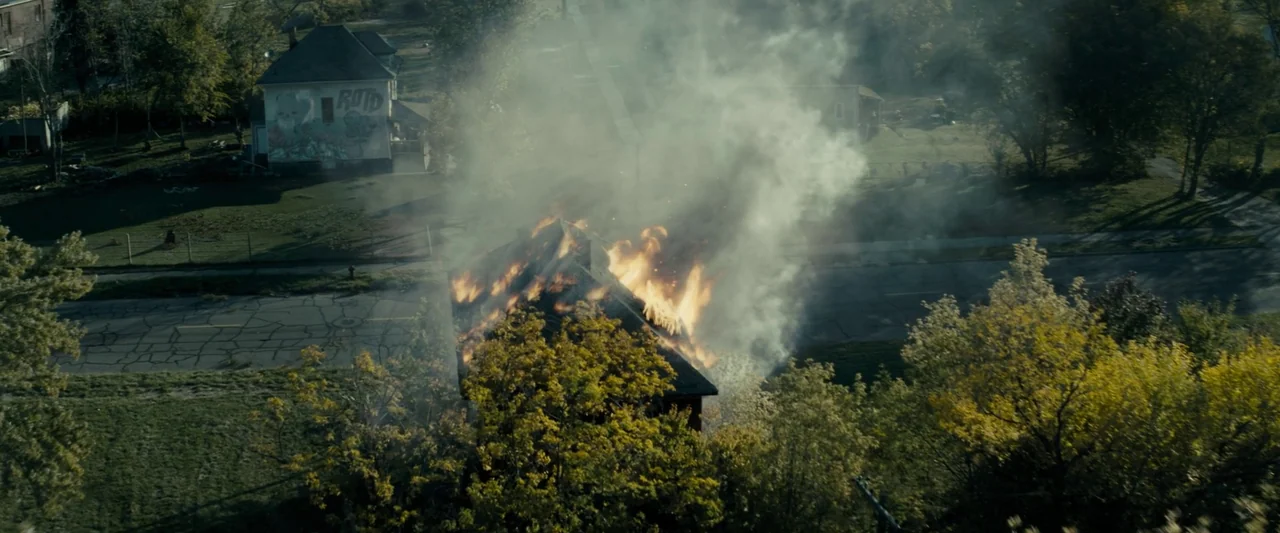 Don’t Breathe 2 (2021) movie still: establishing — An aerial view of a suburban neighborhood shows a small house engulfed in flames. Thick grey…