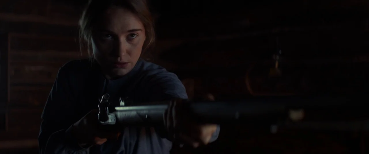 Never Grow Old (2019) movie still: medium — A dark log cabin interior features wooden walls and minimal visible furniture. A young woman holds a…