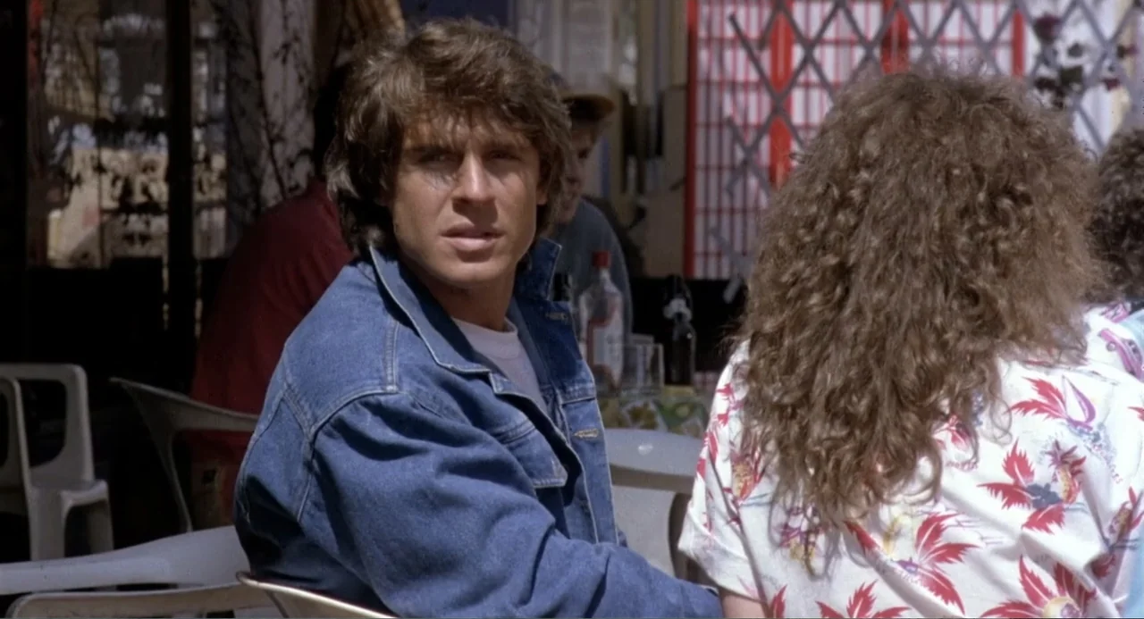 American Ninja 3: Blood Hunt (1989) movie still: medium — An outdoor cafe area has white plastic chairs and a table with bottles. A young man in a…