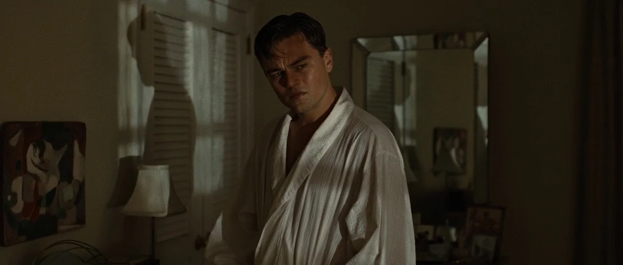 Revolutionary Road (2008) movie still: medium — A dimly lit room with closed louvered doors and a wall-mounted rectangular mirror. A man in a white…